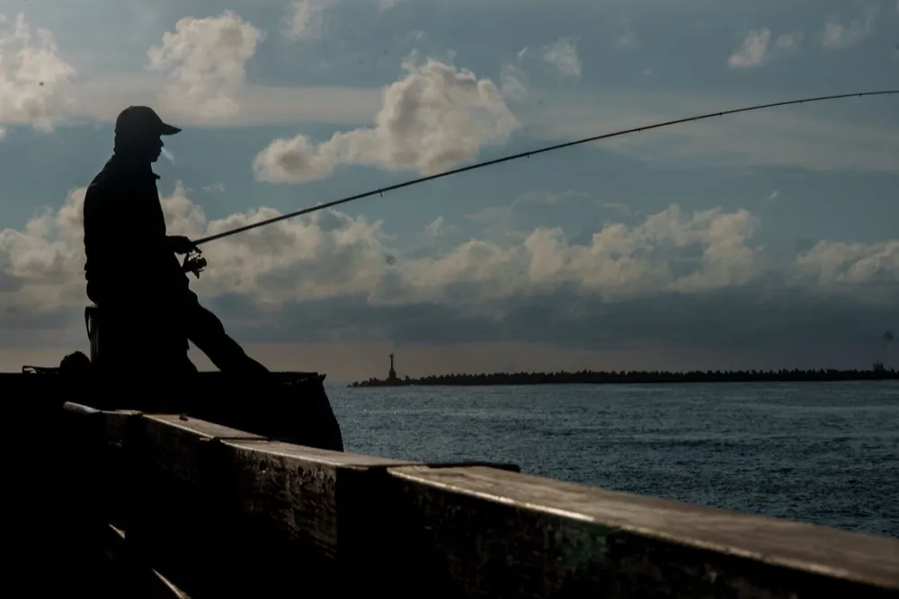 A man fishing off a sea wall, silhouetted against a cloudy sky